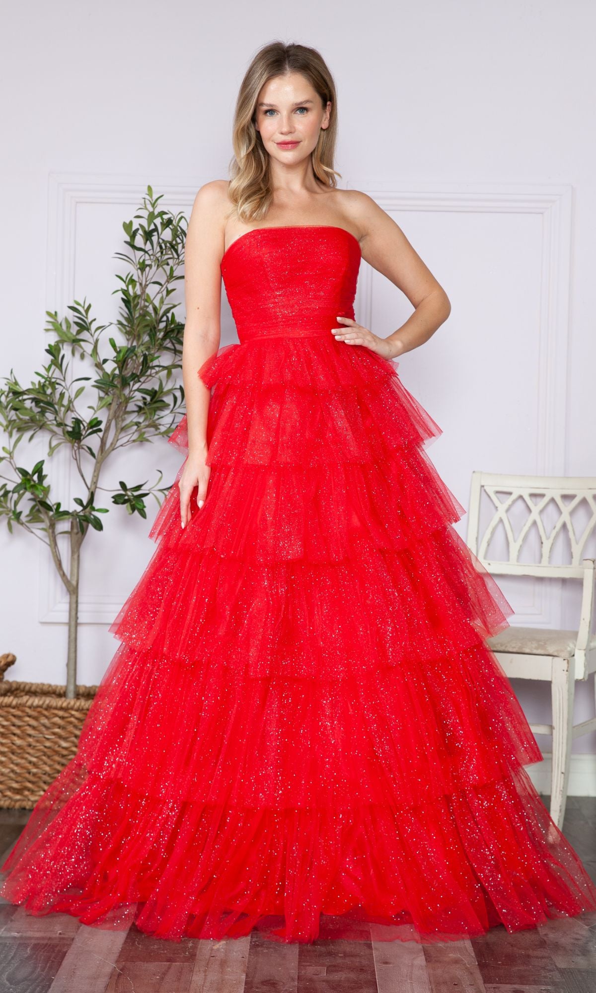 A woman in the Strapless Long Tiered Glitter Prom Ball Gown 9386—an elegant, floor-length red tulle dress with layered ruffles—stands indoors next to a white chair and a potted plant.